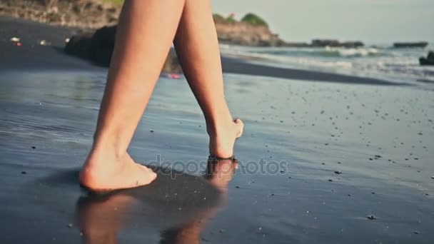 Jovem mulher caminhando ao longo da borda das águas. Uma mulher a percorrer a costa na areia negra. A câmara está a mover-se atrás da fêmea . — Vídeo de Stock