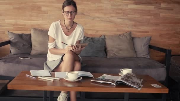 Fiatal nő szemüveg portré kávézóban ült, és a rajz a Jegyzettömbben. Kávét iszik. Kültéri működő fogalom. Kávéház helyet napos loft — Stock videók