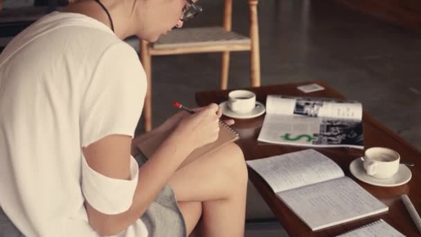 Close-up young woman in glasses portrait sitting in cafe and drawing in notepad. Drinking coffee. Outdoor working concept. Coffee-house space in sunny loft — Stock Video