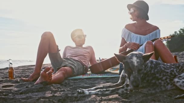 Casal jovem se divertindo na praia. Fim de semana na ilha. Casal jovem cantando com uma guitarra na praia seu cão engraçado. Piquenique à beira-mar — Vídeo de Stock