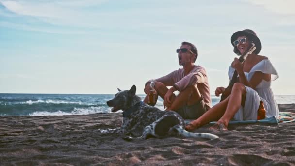 Jong koppel plezier op het strand. Weekend op het eiland. Jong koppel zingen met een gitaar op het strand hun grappige hond. Picknick aan de kust — Stockvideo