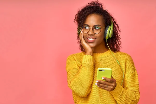 Vreugdevolle zwarte vrouw met tand glimlach, luistert populaire lied via hoofdtelefoon, houdt moderne mobiele telefoon, houdt van luisteren naar muziek tijdens de vrije tijd, geïsoleerd over roze achtergrond. Tieners en technologie — Stockfoto