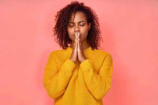 Φωτογραφία της όμορφης νεαρής μαύρης γυναίκας κρατά τα χέρια μαζί σε χειρονομία προσευχής, κλείνει τα μάτια, απολαμβάνει ειρηνική ατμόσφαιρα, έχει ευχάριστη έκφραση, απομονώνονται σε ροζ φόντο. Ελπίζω για το καλύτερο στη ζωή. — Φωτογραφία Αρχείου
