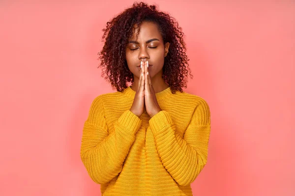 Foto de bela jovem mulher negra mantém as mãos juntas em gesto de oração, fecha os olhos, goza de atmosfera pacífica, tem expressão feliz, isolado sobre fundo rosa. Eu espero por melhor na vida — Fotografia de Stock