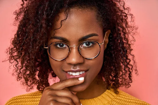 Close up studio shot of beautiful young African American woman model with curly dark hair looking at câmera with a charming cute smile while posing against pink blank, wears fashionable glasses — Fotografia de Stock