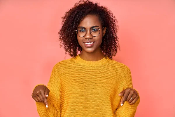 Encantadora mujer negra despreocupada con expresión positiva, apunta hacia abajo con ambos dedos índice, vestida con ropa casual, tiene una amplia sonrisa interesada, aislada sobre fondo rosa. Mira ahí, por favor. — Foto de Stock