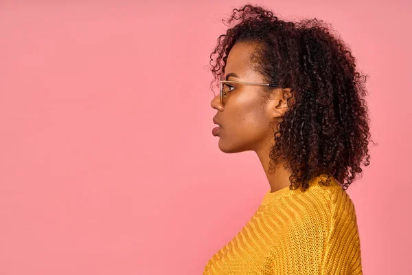 Profil d'une femme afro-américaine sérieuse à la peau pure et saine, porte des lunettes rondes, a une expression contemplative, prête à se promener en plein air, isolée sur un mur de studio rose avec espace de copie — Photo