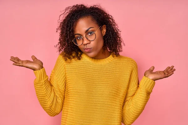 Elszigetelt lövés zavaros gyönyörű afro-amerikai nő, göndör frizura, széttárja a kezét oldalra, vigyorog arc, úgy érzi, kétséges, míg a választás, öltözött alkalmi jumper, elszigetelt rózsaszín fal. — Stock Fotó