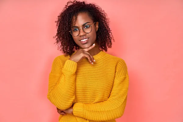 Romántica modelo de piel oscura positiva sonríe alegremente, tiene dientes blancos perfectos, mira directamente a la cámara, tocando su cara, tiene el pelo rizado y tupido, usa suéter naranja. Mujer de aspecto amistoso en pin — Foto de Stock