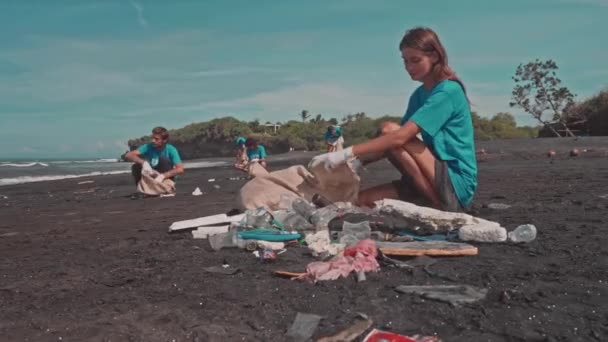 Mujer joven y amigos en camiseta azul recogiendo basura en bolsas ecológicas en la playa — Vídeo de stock