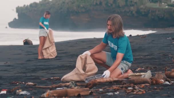 Wolontariuszka w niebieskiej koszulce czyści gruzy plaży, składając śmieci w eko-torbie — Wideo stockowe