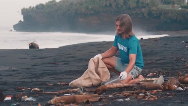 Voluntario en camiseta azul limpia los escombros de una playa, basura plegable en bolsa ecológica — Vídeo de stock