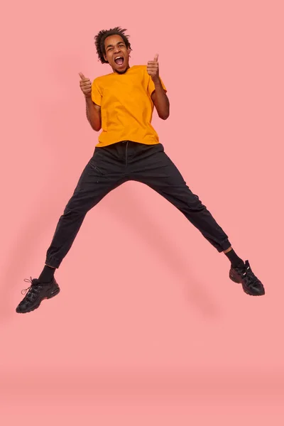 Glad nöjd svart man hoppar i luften och visar med båda händerna godkännande gest, håller två tummar upp, ler brett, bär gul t-shirt, mot rosa vägg. Det är fint, jag gillar det. — Stockfoto