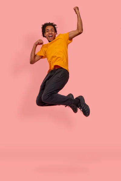 Full längd foto av glada afroamerikanska mannen hoppa upp nävar att skrika ja fira en seger eller glädjas med försäljning start, bära orange casual t-shirt isolerad över rosa bakgrund. — Stockfoto