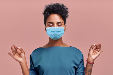 Sakin doktor, solunum maskesi takarak meditasyonu rahatlatıyor, iş konseptinde stresten uzak, düşünceli, genç bayan sağlık görevlisi bej duvarda yoga egzersizleri yapıyor. Coronavirüs.