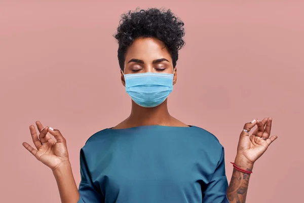 Sakin doktor, solunum maskesi takarak meditasyonu rahatlatıyor, iş konseptinde stresten uzak, düşünceli, genç bayan sağlık görevlisi bej duvarda yoga egzersizleri yapıyor. Coronavirüs.