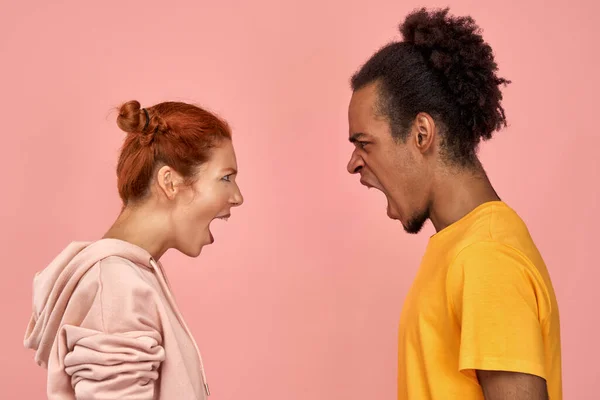 Furious angry black man and ginger woman have dispute, yell loudly at each other, quarrel and sort out relationships, have disagreement, different point of views, stands over pink background. — Stock Photo, Image