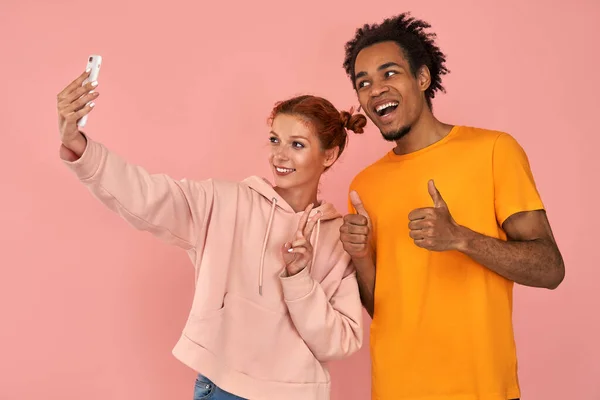 Cheerful black man laughs sincerely while poses for selfie with ginger girlfriend makes peace sign or V gesture, have fun together, shows use modern cell phone for entertaining, on pink background