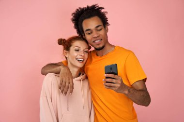 Olumlu genç melez çift birbirlerini kucaklıyor, cep telefonlarına sevinçle bakıyor, sıradan fotoğrafları görmekten memnun, pembe arka plana karşı iyi kablosuz bağlantıdan memnun. İlişki kavramı