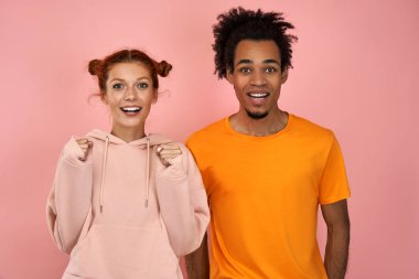 Photo of emotional ginger female and dark skinned male clench fists, exclaim and support favorite sports team, have overjoyed face expressions, dressed in casual wear, isolated on pink background clipart