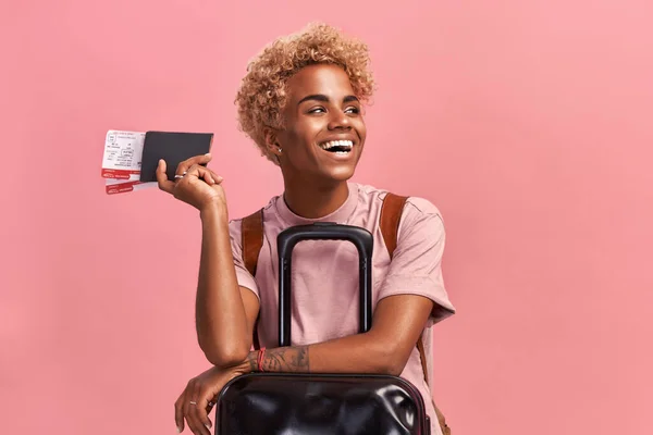 Indoor shot van optimistische Afro vrouwelijke toerist met bagage houdt paspoort en tickets, wacht op de vlucht, klaar voor vakantie, geniet van reizen, ontvangt visum voor reizen naar het buitenland, gekleed in casual outfit — Stockfoto