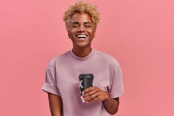 Feliz optimista mujer de piel oscura gestos activamente, bebidas calientes, habla emocionalmente con amigos, usa camiseta lila, tiene buena taza de café aumenta la energía con cafeína, sobre fondo rosa — Foto de Stock