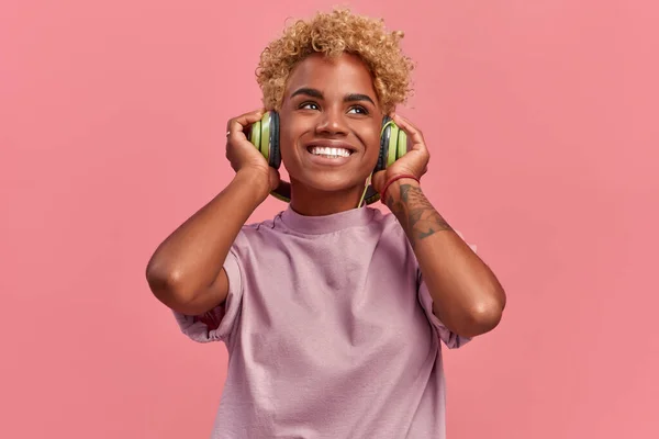Glad ung afroamerikansk kvinna avslappnad med en bra låt har ett brett leende, berör hörlurar, lyssnar på musik, klädd i en lila casual t shirt, isolerad över rosa studio vägg, vill dansa — Stockfoto