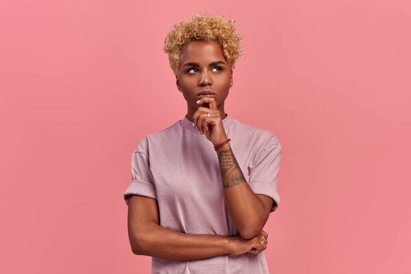 Pensive attractive curly blonde African American female being deep in thoughts, looking up, keeps forefinger near the chin, wears fashionable lilac t shirt, stands against a pink background