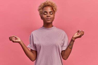 Sakin, rahatlamış, koyu tenli bir kadın derin bir nefes alır, Zen jestinde ellerini yana tutar, Nirvana 'ya uzanır ve yoga yapar, gözleri kapalı durur, stresten uzak, pembe arka planda durur.