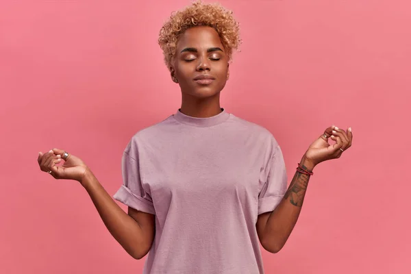 Calm relieved blondish dark skinned woman takes deep breath, keeps hands sideways in zen gesture, reaches nirvana and practices yoga, stands with closed eyes, stress free, against pink background — Stock Photo, Image