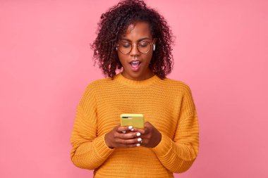 Çekici kıvırcık kıvırcık saçlı, Afro saçlı kadın akıllı telefon ekranında tedirgin görünüyor. Arkadaşından gelen mesajdan etkilenmiş. Pembe arka planda yüz ifadesine şaşırmış.