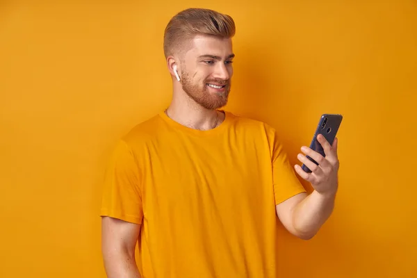 Cheerful, friendly-looking handsome redbeard caucasian man talking on phone, answer with tapping wireless earphone, wearing headphone communicating using modern device isolation over orange background