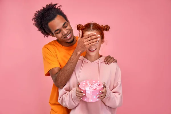 Jóképű afro-amerikai férfi fedezi a boldog vörös hajú kaukázusi barátnő szemét, és így neki jelen kis doboz, visel narancs alkalmi póló és rózsaszín kapucnis, elszigeteltség rózsaszín háttér — Stock Fotó