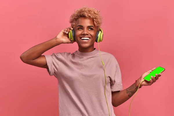 Overjoyed dark skinned girl stays alive with sound of music, keeps hands on headphones, has modern gadgets, listens song and feels glad, expresses happiness. Curly woman enjoys audio book, pink wall