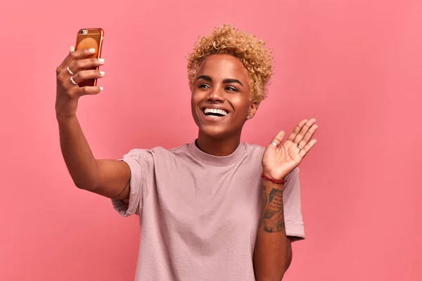 Glad mörkhyad glad kvinna med blond afro frisyr, håller modern mobiltelefon framför ansiktet, vågor palm i kameran, gör videosamtal, klädd i casual wear, isolerade över rosa bakgrund — Stockfoto
