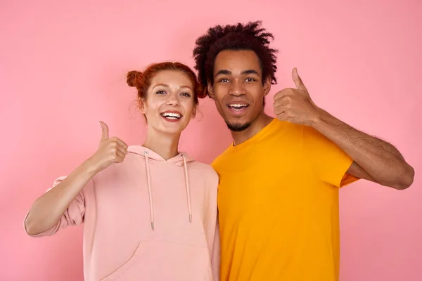 Portré elégedett szép romantikus multikulturális pár, gyömbér nő és afro-amerikai férfi mutatja hüvelykujj fel jelzi értékesítési kedvezmények úgy érzi, izgatott viselni modern ruhát elszigetelt rózsaszín háttér — Stock Fotó