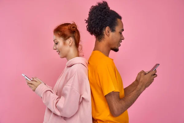 Beautiful, pretty, cute red hair woman and handsome dark skinned man standing back to back and writing messages through internet on modern smartphones, enjoy nice conversation over pink background