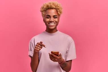 Neşeli, gülümseyen Afrikalı Amerikalı kadın, cep telefonunun ekranına işaret ediyor, yeni bir uygulaması olduğu için seviniyor, online iletişimden hoşlanıyor, yüksek bir ruh hali içinde, pembe arka planda izole edilmiş. Teknoloji