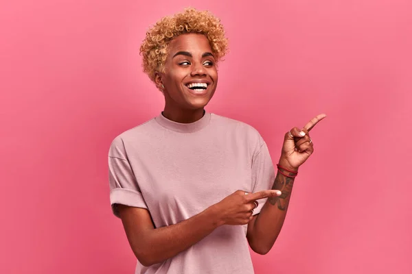 Prise de vue horizontale femme à la peau foncée et aux cheveux blonds afro, pointe les deux index, montre un espace vide pour votre promotion, isolée sur fond rose. Personnes, concept publicitaire — Photo