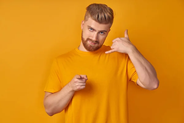 Call me back please. Young attractive blonde guy with red beard, makes phone sign, indicates with index finger directly at camera, wants to speak with someone via cell phone isolated orange background — Stock Photo, Image