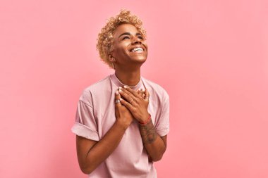 Dokunulmuş güzel, gülümseyen Afro-Amerikalı kız avuçlarını kalbinde tutuyor. Minnettarlığını, memnuniyetini ve minnettarlığını ifade ediyor. Pembe arka planda bir lavanta tişörtü giyiyor. Aşk konseptinde itiraf.