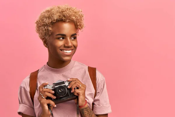 Obrázek talentované Afro fotografky pořizuje profesionální záběry během dobrodružné cesty, používá retro kameru, nosí levandulové tričko a batoh, užívá si dovolenou, pózuje v interiéru. Koncept aktivního odpočinku — Stock fotografie