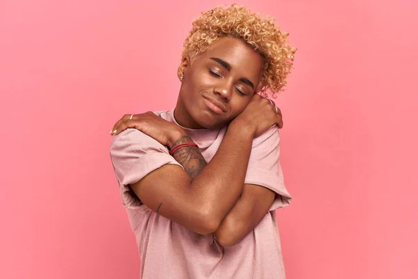 Charmig vacker afro kvinna med nöjda känslor, kramar sig själv, håller ögonen stängda, ler med glädje, känns komfort, bär lavendel t-shirt, lutar huvudet, har hög självkänsla över rosa bakgrund — Stockfoto