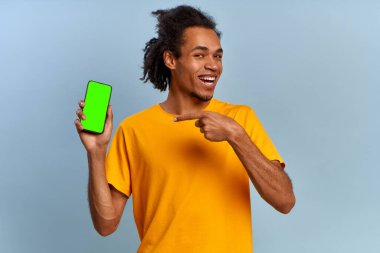 Gülen genç Afrikalı Amerikalı hippi, mavi arka planda izole edilmiş alfa kanallı modern akıllı telefon yeşil ekranını işaret ediyor. Yeşil ekran reklam alanı. Araçlar ve iletişim konsepti
