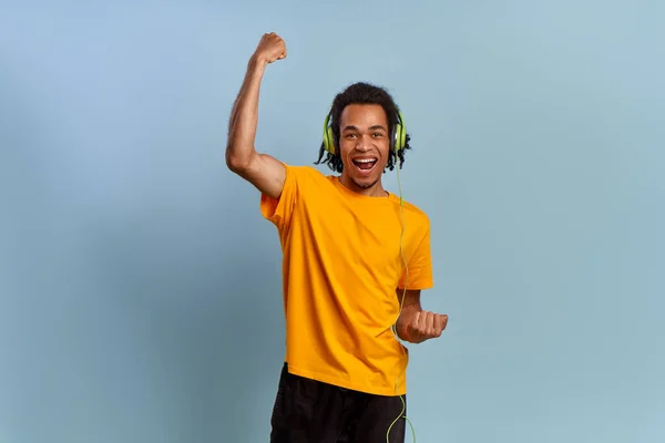 Glädjande och ung afrikansk amerikansk kille med hörlurar skildrar glädje, känner sig glad och överlycklig, lyfter knutna nävar, njuter av bra låtvibbar på blå bakgrund. Begreppet framgång och vinnande lotteri — Stockfoto