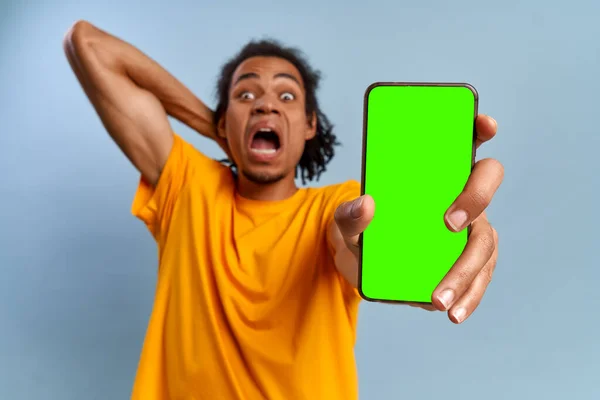 Følelsesmæssige chokeret afrikansk amerikansk ung mand viser grøn skærm moderne smartphone med alfa-kanal på blå baggrund. Begrebet rabatter og kampagner i online butik. Reklameplads - Stock-foto