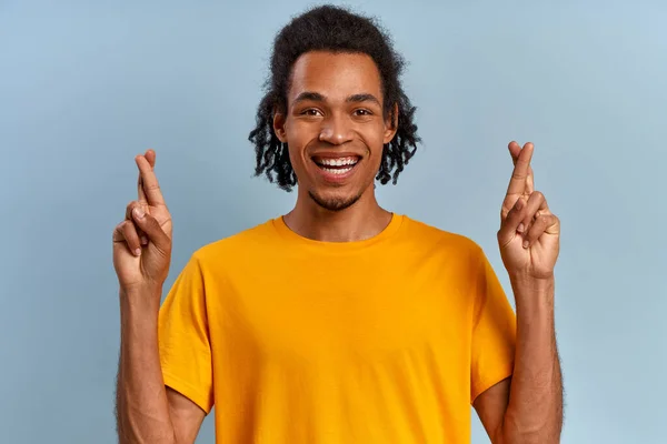 Leende afrikansk student kille i gul t-shirt korsade fingrarna på händerna på blå bakgrund. Begreppet fans för fotboll laget och önskan om en bra examen. Reklamutrymme — Stockfoto