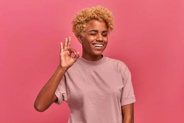 Sorrindo linda mulher americana africana com cabelos loiros diz bom trabalho ou bem feito, faz gesto bem, demonstra símbolo de aprovação e como, pisca os olhos sobre fundo estúdio rosa — Fotografia de Stock