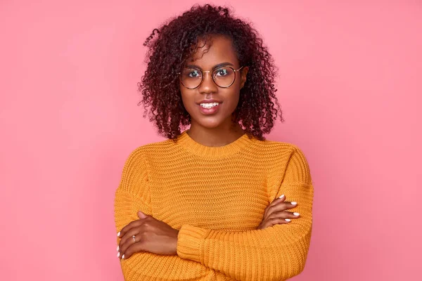 Cute african American joven hipster hembra en un jersey amarillo con sus brazos cruzados sobre fondo rosado en estudio. Concepto de una mujer joven exitosa. Lugares para la publicidad — Foto de Stock