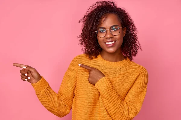 Una jovencita afriquísima y alegre estudiante afriamericana de dulzura amarilla señala a la izquierda basándose en el fondo rosa del estudio. Concepto de publicidad de productos juveniles. Copie el espacio para su texto. — Foto de Stock